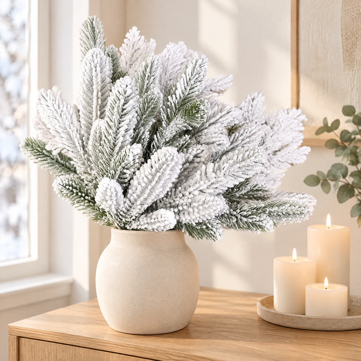 Snowy flocked pine picks in a cream ceramic vase on an oak sideboard with candles