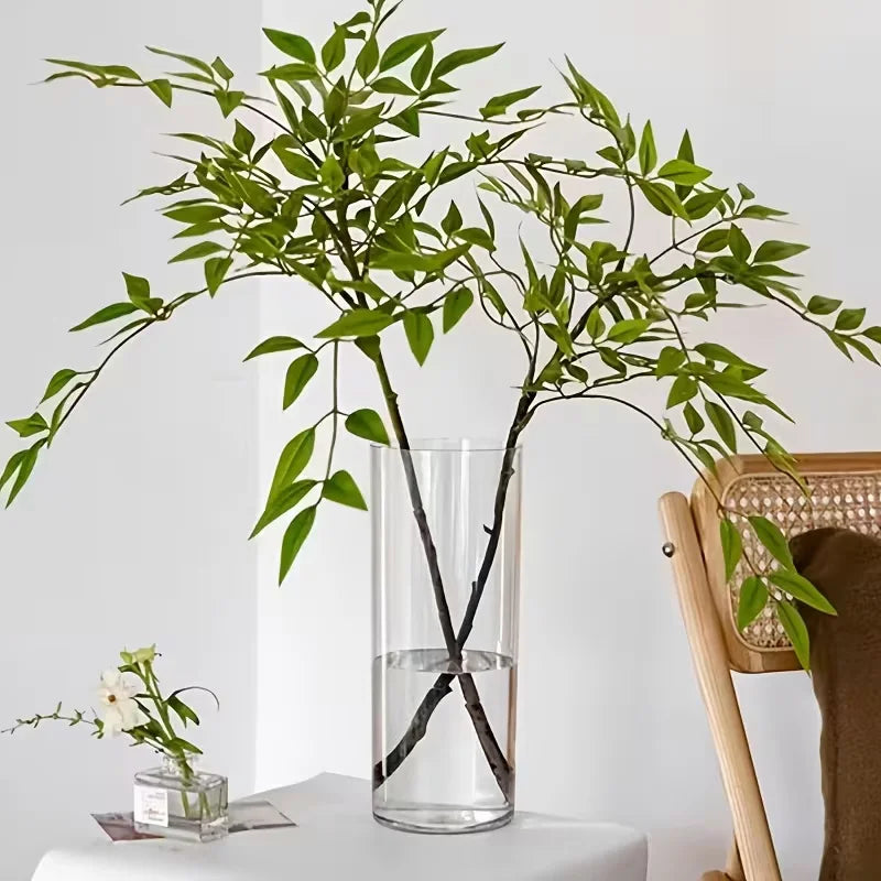 Clear acrylic floor vase with tall green branches next to a white wall and a wicker chair