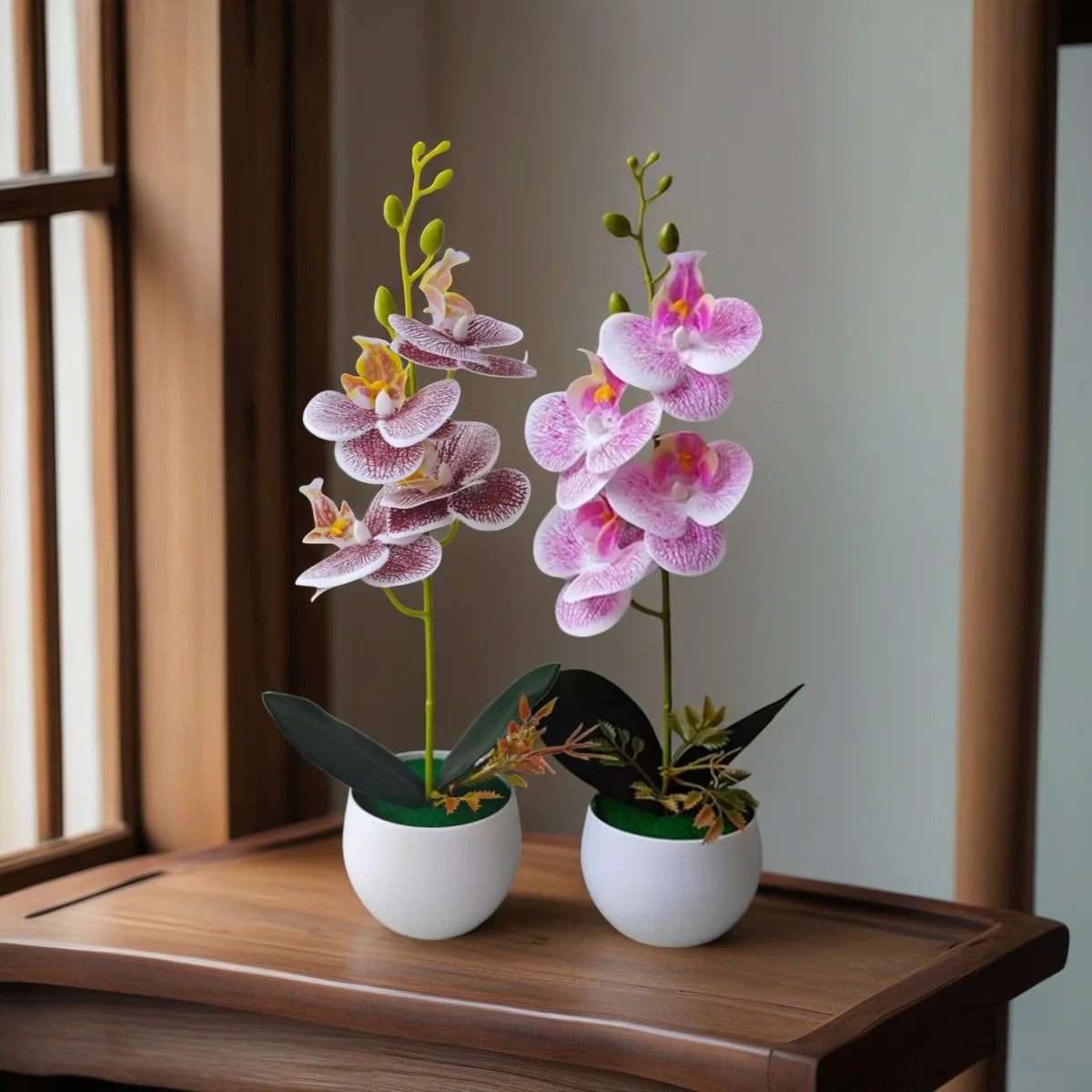 Two artificial pink orchids in white pots on a wooden table by a window