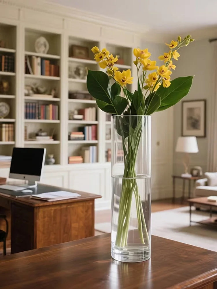 Tall clear cylinder vase with yellow flowers in a bright living room with bookshelves